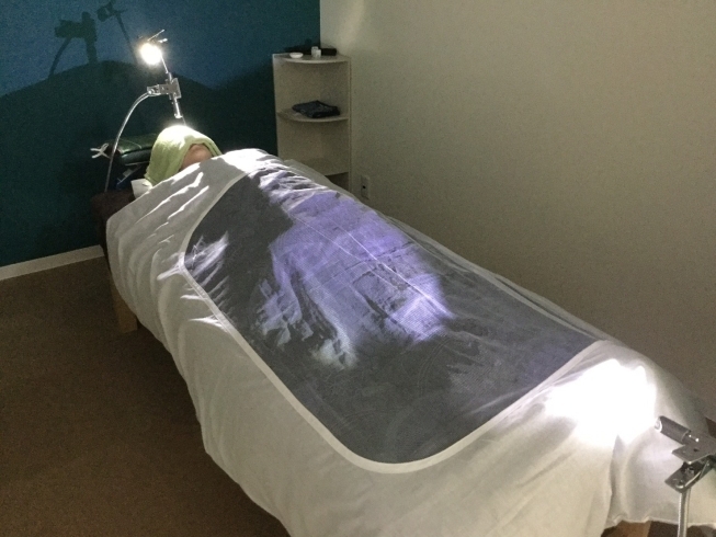 Inside the room it is possible to see a patient lying in bed and covered with a blanket and there is a light that radiates luminous ions onto the patient's head.