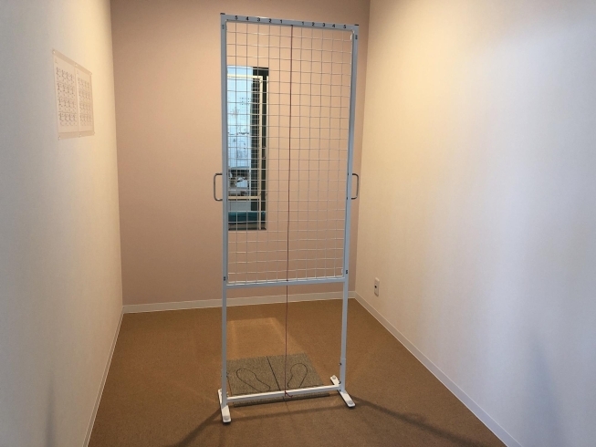 In a room there is a white grid that is the average height of an adult, and in the middle there is a red cord stretched vertically to the white grid and in the background you see a long mirror.