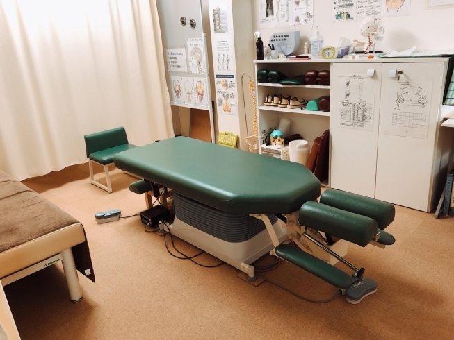 This is an automatic stretcher that is vertically adjustable and allows the chiropractor to manipulate the height appropriate for the patient's comfort. Next to the stretcher is another fixed-height stretcher and in the back is a green upholstered chair.